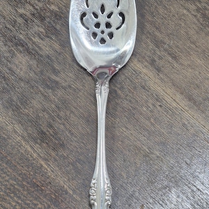 Könnte beinhalten: Silberner Tortenheber mit dekorativem Griff und einer geschlitzten, ovalen Klinge. Die Klinge hat ein florales Ausschnittmuster. Der Griff hat ein verziertes, geprägtes Muster. Der Heber besteht aus glänzendem, reflektierendem Metall.