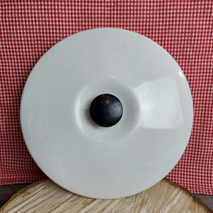 Peut inclure: Un couvercle en céramique blanc avec un bouton noir au centre. Le couvercle est rond et présente un design légèrement concave. Le fond est un motif à carreaux rouges et blancs.