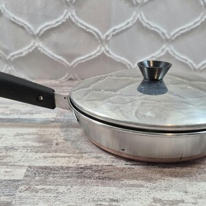 May include: A stainless steel frying pan with a matching lid and a black handle. The handle features a small metal loop at the end for hanging. The pan has a polished metallic finish, a conical black knob on the lid, and a visible copper-coloured base.