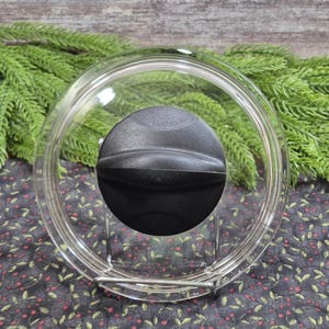 May include: A circular, transparent glass lid for cookware featuring a central, black, rounded plastic knob. The lid has a raised metal rim around the outer edge and is positioned upright on a small, silver-toned wire display stand.