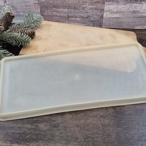 May include: A rectangular, semi-transparent, off-white plastic lid with a raised border edge. The lid features a smooth, flat surface and is designed to fit a container, showcasing a simple, functional, and utilitarian construction.
