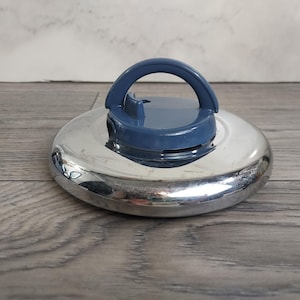 Puede incluir: Una tapa de acero inoxidable con un asa y una parte superior de plástico azul. La tapa es redonda y reflectante, con un asa azul que se arquea sobre la parte superior. El asa es una pieza sólida y curvada, y la parte superior tiene una pequeña sección elevada.