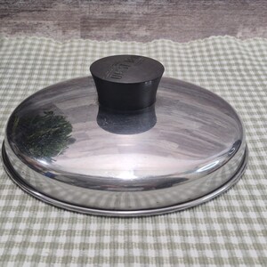 Peut inclure: Un couvercle de casserole en acier inoxydable avec une poignée noire. Le couvercle est rond et réfléchissant, avec une forme légèrement bombée. La poignée est cylindrique et porte du texte. Le couvercle est posé sur une surface à carreaux.