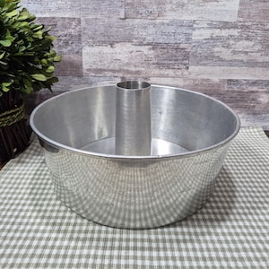 May include: A silver-colored angel food cake pan with a central tube. The pan is round with a wide base and a slightly flared rim. It sits on a green and white checkered cloth.