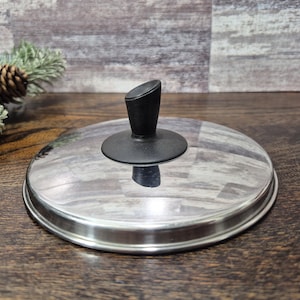 May include: A stainless steel and glass lid with a black handle. The lid is round with a shiny, reflective surface. The handle is angled and made of black plastic. The lid is designed for cookware.