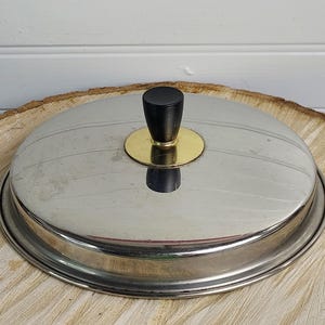 Può includere: Un piatto da portata in acciaio inossidabile con pomello nero e accento dorato. Il coperchio è rotondo con un design a coste. Il piatto poggia su una superficie di legno.