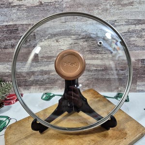 Op de afbeelding: Een heldere glazen T-fal deksel met een koperkleurige handgreep en een roestvrijstalen rand. Het deksel wordt getoond op een zwarte standaard, rustend op een houten snijplank. De achtergrond is een rustieke houten muur.