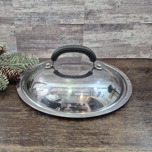 Puede incluir: Una tapa de olla circular con un centro de vidrio transparente y un borde de acero inoxidable pulido. La tapa incluye un asa negra arqueada fijada de forma segura en la parte superior con dos remaches metálicos para facilitar su manejo.