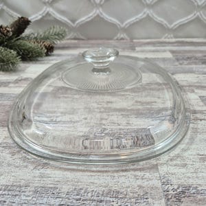 May include: A clear glass lid with an oval shape and a central knob handle. The top surface features a circular, radiating sunburst pattern surrounding the knob, while the outer edge of the lid is smooth and transparent, designed for a dish.