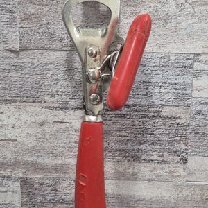 May include: A vintage can opener with a bottle opener. The opener is made of silver metal with a red handle and a red lever. The handle has the numbers "00" on it. The can opener is on a wooden surface.
