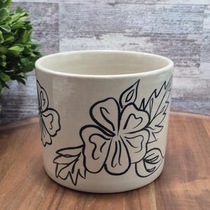 May include: A cream ceramic plant pot with a black floral design. The cylindrical pot has a wide opening and is decorated with hand-drawn flowers and leaves. The design wraps around the exterior.