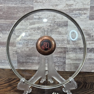 Könnte beinhalten: Ein klarer Glasdeckel mit einem Edelstahlrand und einem kupferfarbenen Griff. Der Griff trägt die Buchstaben "PD" in einer dekorativen Schriftart. Der Deckel wird auf einem transparenten Kunststoffständer präsentiert.