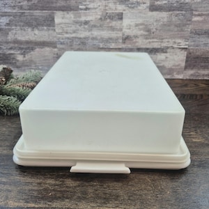 May include: A rectangular, off-white plastic storage container with a matching lid. The container features a smooth, solid surface and a base with a small tab handle on the front edge, designed for secure closure and easy opening of the storage unit.