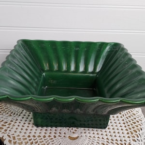 Może przedstawiać: Kwadratowa, zielona ceramiczna miska z falistą krawędzią i teksturowaną podstawą. Misa ma żebrowany wzór po bokach i stoi na białej koronkowej serwetce. Misa jest pusta.