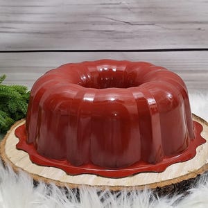 May include: A glossy, deep red bundt cake, presented on a rustic wooden slice. The cake has a classic ring shape with vertical ridges. The wooden slice sits on a bed of white faux fur, with a sprig of green foliage in the background.