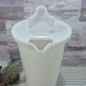 May include: A white plastic pitcher with a hinged lid. The pitcher has a spout for pouring and is designed for liquids. The lid is open, revealing the interior. The pitcher is on a checkered surface.