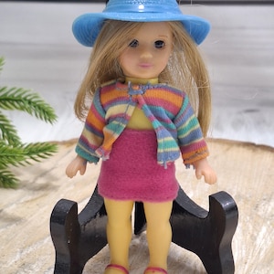 May include: A doll wearing a light blue hat, a striped sweater, and a pink skirt. The doll has blonde hair and is standing on a black stand. The doll is wearing yellow shoes with pink straps.