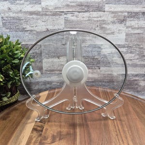 Peut inclure: Couvercle de casserole en verre transparent avec un bord en acier inoxydable et un bouton blanc. Le couvercle est présenté sur un support en plastique transparent. Le diamètre du couvercle est d'environ 25 cm.