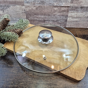 May include: A circular clear glass pot lid featuring a polished stainless steel rim and a matching stainless steel knob handle centered on top. The lid includes a small circular steam vent hole positioned near the lower edge of the glass surface.