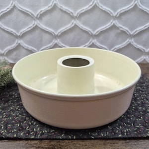 May include: A round, cream-colored ceramic tube pan with a smooth interior and a central chimney. The exterior features a subtle, textured finish, and the top rim is slightly rounded. The pan has a wide, open basin design for baking cakes.