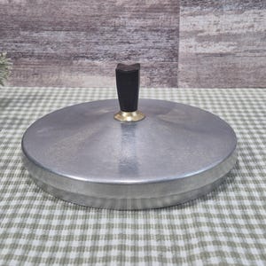 May include: A round, silver-toned metal lid with a black handle and gold-colored base. The lid is sitting on a green and white checkered surface. The lid is likely for a pot or pan.