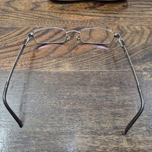 K&ouml;nnte beinhalten: Eine rechteckige Brille mit einem d&uuml;nnen silbernen Metallrahmen und dunkelgrauen B&uuml;geln. Die Brille verf&uuml;gt &uuml;ber klare Gl&auml;ser, verstellbare Nasenpads und einen Steg, der die beiden Brillengl&auml;ser verbindet, gesehen von der R&uuml;ckseite.
