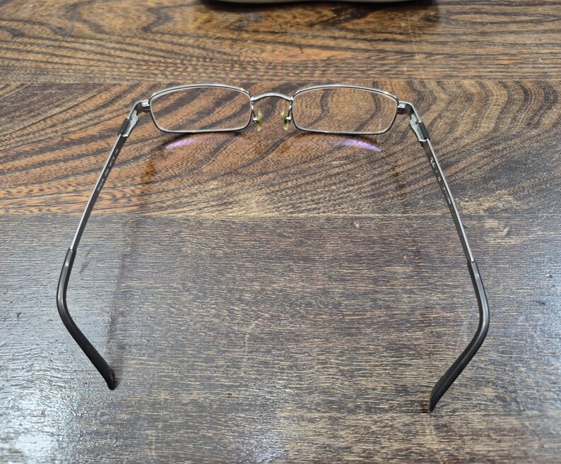 K&ouml;nnte beinhalten: Eine rechteckige Brille mit einem d&uuml;nnen silbernen Metallrahmen und dunkelgrauen B&uuml;geln. Die Brille verf&uuml;gt &uuml;ber klare Gl&auml;ser, verstellbare Nasenpads und einen Steg, der die beiden Brillengl&auml;ser verbindet, gesehen von der R&uuml;ckseite.
