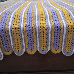 Puede incluir: Una manta tejida a mano con rayas verticales en color amarillo mostaza, lavanda y blanco. La manta presenta un borde festoneado con colores alternos, creando un patrón texturizado y repetitivo en toda la superficie de la tela.
