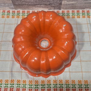 May include: An orange, fluted bundt cake pan with a central hole. The vintage metal pan has a glossy finish with some wear and tear. The pan is sitting on a floral patterned tablecloth.