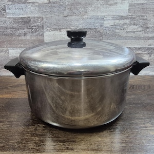 May include: A stainless steel cooking pot with a lid and black handles. The pot has a shiny, metallic finish and a black knob on the lid. The pot is sitting on a wooden surface.
