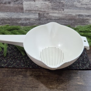 May include: A white plastic colander with a handle and a perforated base. The colander is set on a dark, patterned surface, with green foliage in the background. The colander is designed for straining liquids from food.