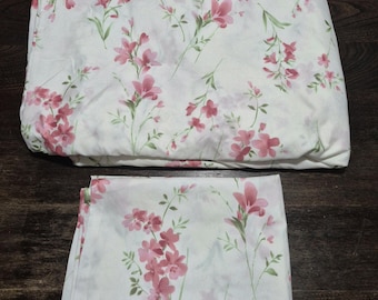 Puede incluir: Juego de cama blanco con un diseño floral en tonos rosa y verde. El juego incluye una sábana doblada y una sábana plana, ambas con el mismo diseño botánico repetitivo. Las sábanas están hechas de una tela suave y ligera.