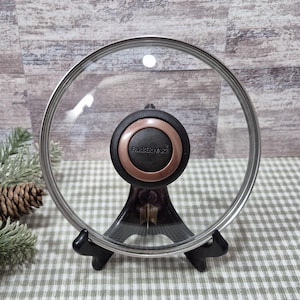 Puede incluir: Una tapa de cristal transparente Farberware con borde de acero inoxidable y un asa negra y cobriza. La tapa se exhibe sobre un soporte negro contra un fondo de pared de madera y un mantel a cuadros verdes y blancos.