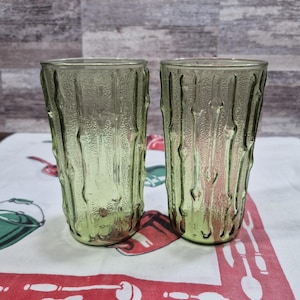 May include: Two green-tinted drinking glasses with a textured, bark-like design. The glasses are tall and cylindrical, with a slightly flared top. The textured surface gives them a unique, rustic appearance. The glasses are empty and sit on a patterned tablecloth.
