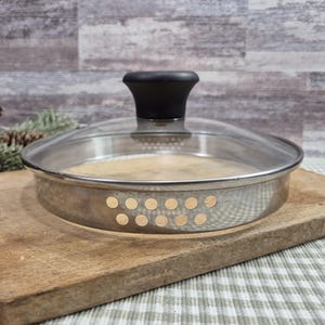 Può includere: Un cestello per cottura a vapore in acciaio inossidabile con coperchio in vetro trasparente e manico nero. Il cestello ha un design perforato con piccoli fori ed è appoggiato su un tagliere di legno.