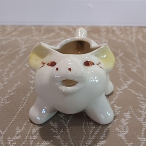 May include: A ceramic pig-shaped planter. The planter is white with yellow accents on the ears and brown details for the eyes and snout. It has a small opening at the top for planting. The planter is designed to look like a pig.