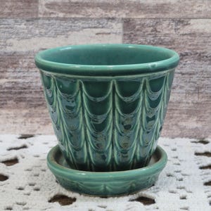 Puede incluir: Una maceta de cerámica color turquesa con un platillo a juego. La maceta tiene un patrón texturizado y repetitivo en el exterior. Es redonda con una abertura ancha. El platillo es un plato poco profundo.