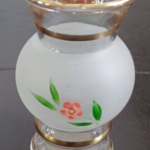 May include: A small, clear glass vase with a frosted, bulbous body and a clear top. The vase is decorated with a gold rim and a painted floral design. The vase is approximately 5 inches tall.