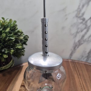May include: A vintage glass jar with a metal lid and a plunger mechanism. The plunger has a dark red, round handle. The jar is clear, and the mechanism is silver-coloured. The jar sits on a wooden surface.