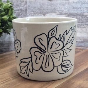 May include: A cylindrical ceramic planter with a smooth off-white finish, featuring a hand-drawn black line art design of a large hibiscus-style flower with detailed leaves wrapping around the exterior surface of the vessel.