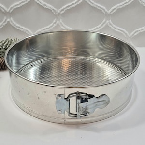 May include: A round silver-colored metal springform cake pan with a textured base and a side latch mechanism. The pan features a smooth exterior wall and a dimpled interior surface, designed for baking cakes with a removable outer ring.