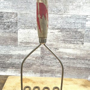 Puede incluir: Pasapurés vintage con mango de madera con detalles de pintura roja. El cabezal de metal para triturar tiene un diseño ondulado. Un utensilio de cocina para triturar patatas y otras verduras. El mango tiene un agujero para colgar.