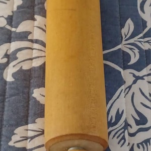 May include: A wooden rolling pin, a kitchen utensil, with a cylindrical body and two handles. The rolling pin is made of light-coloured wood and is on a blue and white floral patterned surface.