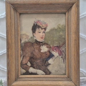 May include: A framed print featuring a portrait of a person wearing a brown high-neck garment, white gloves, and a floral hat, holding a dark umbrella. The artwork is enclosed in a rectangular, medium-toned wooden frame with a grooved border.