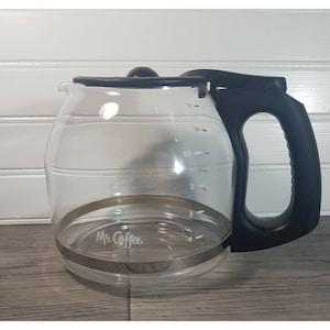 Puede incluir: Una jarra de café de cristal transparente con asa y tapa de plástico negro. La jarra presenta marcas de medición impresas en blanco para 4, 6, 8, 10 y 12 tazas, junto con el logotipo blanco de Mr. Coffee impreso en el cuerpo de cristal.