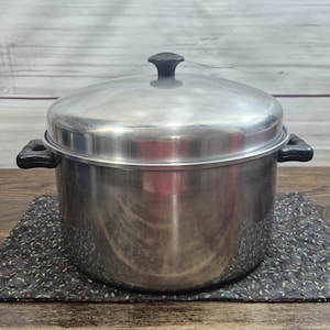 May include: A stainless steel steamer pot with a lid and black handles. The pot sits on a dark floral patterned trivet. The lid has a black knob. The pot is on a wooden surface with a light wood background.