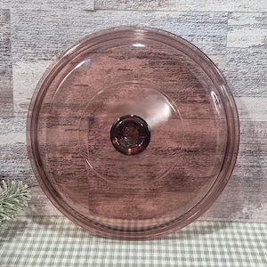 May include: A clear, round, glass lid with a dark purple tint. The lid has a central knob and a slightly raised rim. The background is a wooden surface with a green and white checkered pattern.
