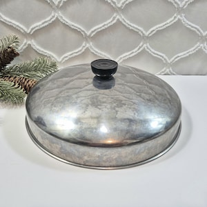 May include: A round, silver-coloured metal lid with a hammered texture and a small, black, circular knob handle centred on top. The lid has a smooth, reflective surface and a slightly domed shape, designed to fit over a circular cooking vessel.