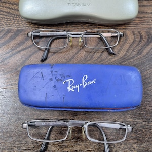 K&ouml;nnte beinhalten: Zwei Paar rechteckige Brillen mit Metallrahmen und schwarzen B&uuml;gelenden sowie zwei Schutzetuis. Ein Etui ist hellgrau mit dem Text Ray-Ban TITANIUM, das andere ist blau mit dem aufgedruckten wei&szlig;en Ray-Ban-Logo auf der Vorderseite.