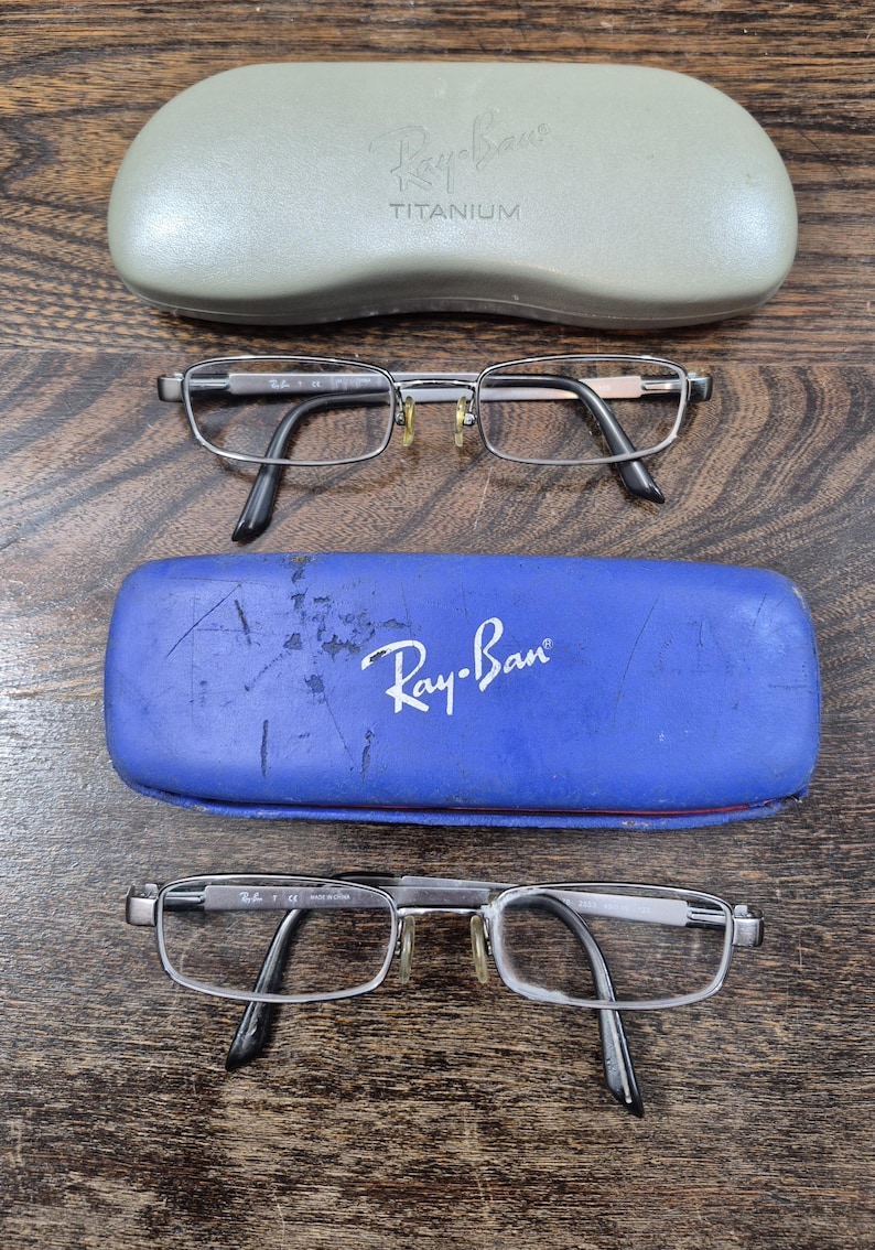 K&ouml;nnte beinhalten: Zwei Paar rechteckige Brillen mit Metallrahmen und schwarzen B&uuml;gelenden sowie zwei Schutzetuis. Ein Etui ist hellgrau mit dem Text Ray-Ban TITANIUM, das andere ist blau mit dem aufgedruckten wei&szlig;en Ray-Ban-Logo auf der Vorderseite.